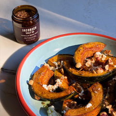 A jar of Jacobsen Salt Co. vanilla bean salt sits next to a light blue plate with a red rim, filled with roasted quash halves topped with nuts and crumbled white cheese.